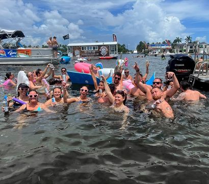 Cheering group of friends partying in shallow coastal water near anchored boats and pontoons, raising drinks and colorful inflatables under a sunny palm-lined waterfront.