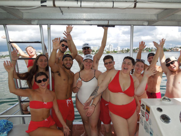 Group of friends in red swimsuits cheering and waving aboard a party boat, bride and groom caps visible, city waterfront in the background.