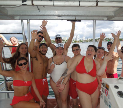 Group of friends in red swimsuits cheering and waving aboard a party boat, bride and groom caps visible, city waterfront in the background.