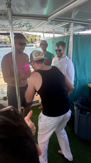 Group of friends partying on a pontoon boat in a sunny marina, passing a large pink funnel drink with waterfront homes and palm trees in the background.