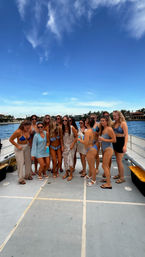Group of women in swimsuits and cover-ups enjoying a sunny yacht party, holding drinks on deck near a palm-lined waterfront with mansions under a bright blue sky.
