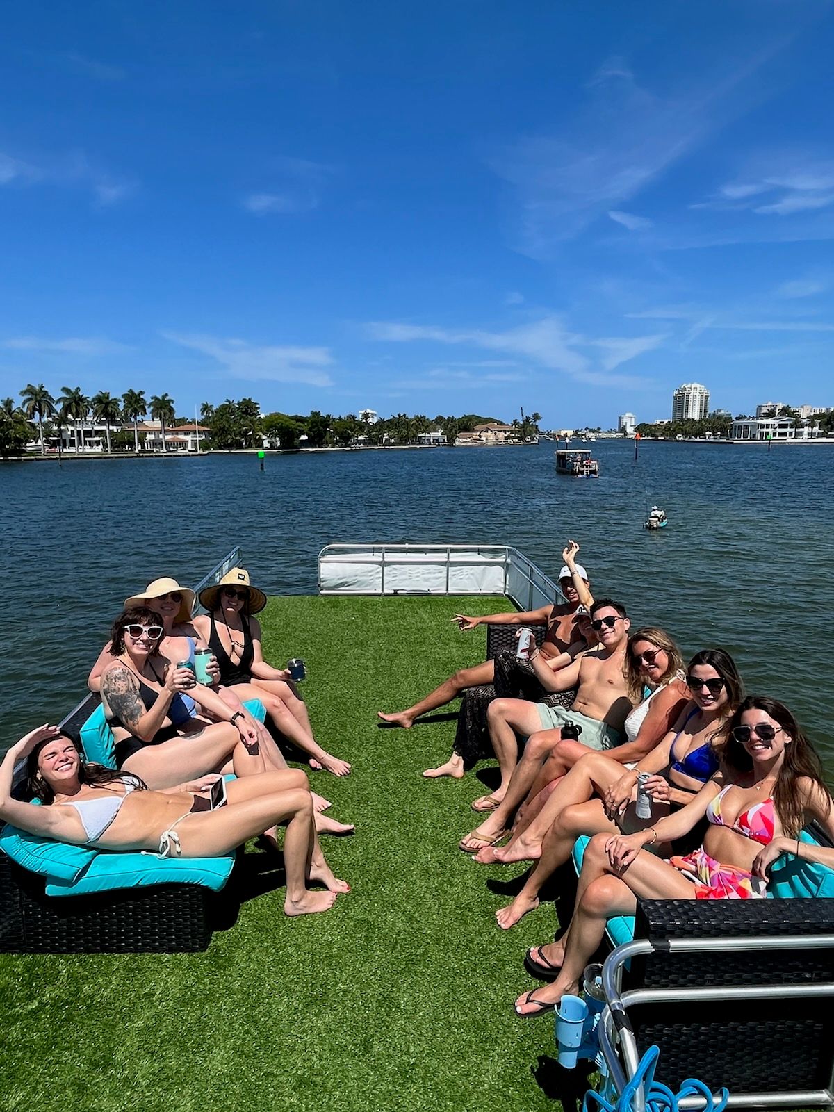 Group of friends sunbathing on a pontoon boat’s grassy top deck, sipping drinks under a bright blue sky with palm‑lined waterfront and distant buildings.