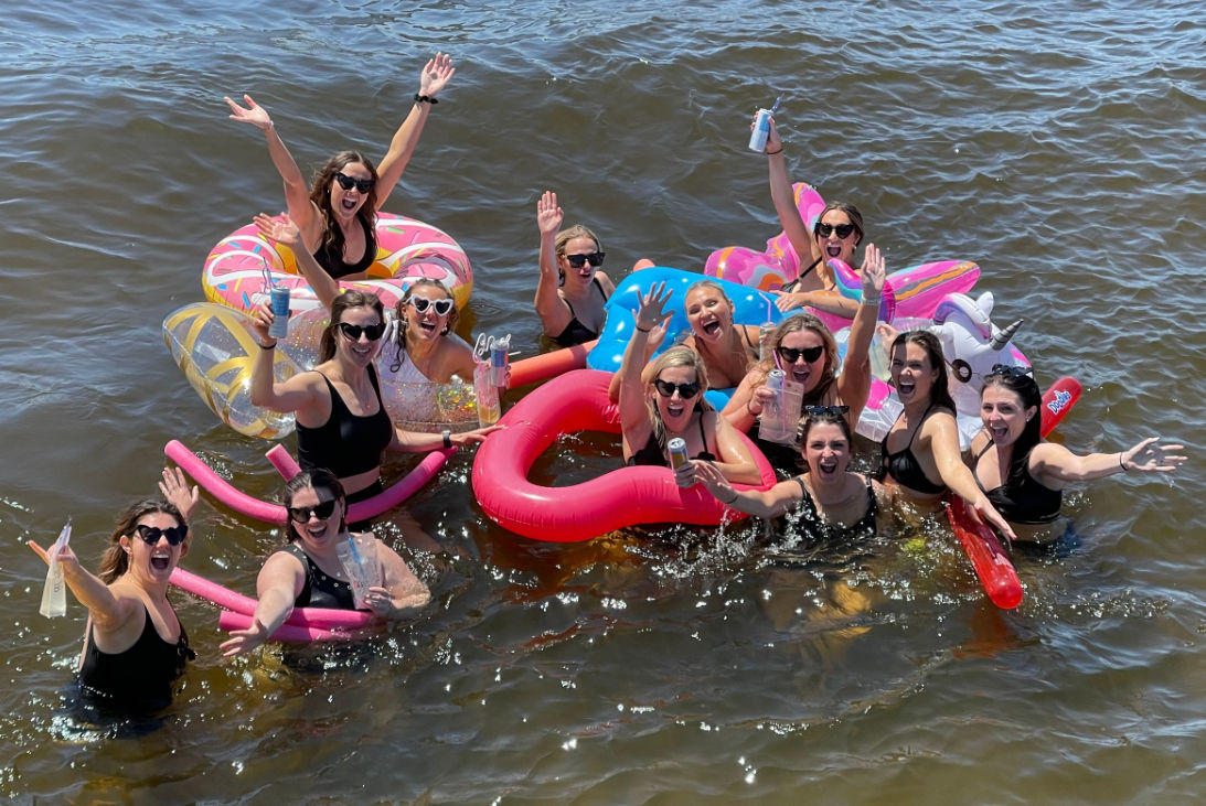 Group of women in swimsuits enjoying a sunny day in open water on colorful inflatable floats (unicorn, donut, heart), waving and holding drinks.
