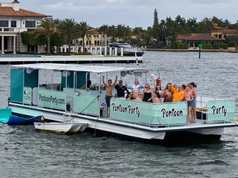 Group of people partying on a mint-green pontoon boat cruising a palm-lined waterfront in front of luxury homes
