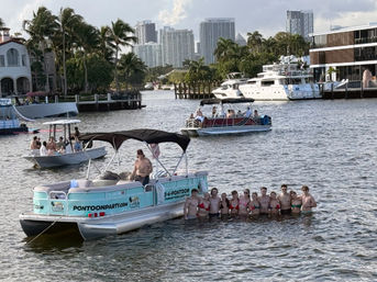 Group of friends waist‑deep in a sunny coastal canal next to a teal party pontoon boat, palm trees, yachts and city skyline in the background.
