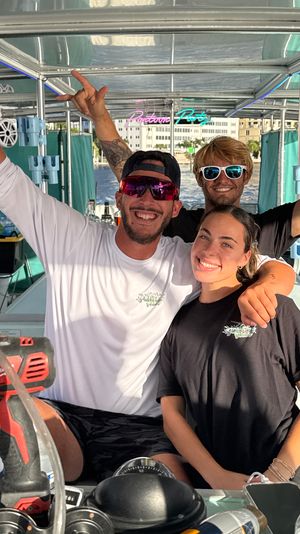 Three smiling friends posing on a pontoon boat at a sunny waterfront marina, wearing sunglasses and casual tees with raised arms and a city skyline in the background.