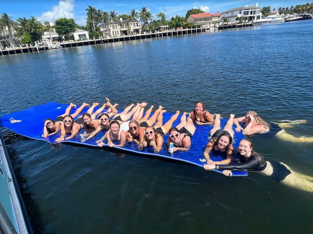 Group of friends lounging on a large blue floating mat in a sunny waterfront canal lined with palm trees and upscale homes, smiling and holding drinks.