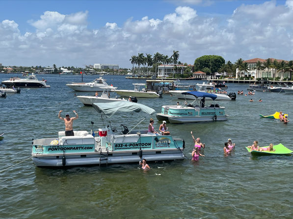 Sunny coastal bay with pontoon boats and pleasure craft, people swimming and lounging on inflatables for a boat‑party vibe, palm‑lined waterfront and luxury homes
