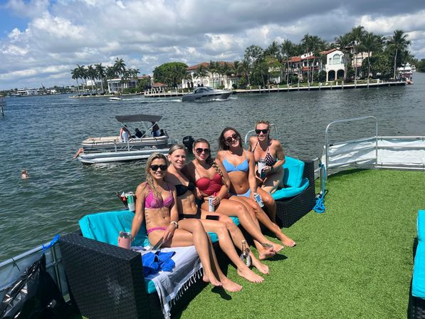 Five smiling women in bikinis lounging on a turquoise-cushioned pontoon boat with artificial turf, holding drinks, with palm-lined waterfront mansions and a partly cloudy sky in the background.