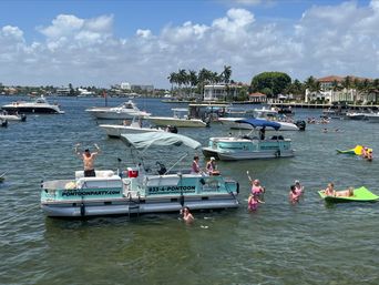 Sunny South Florida intracoastal scene with multiple pontoon boats, people swimming and floating, and waterfront mansions lined with palm trees under a blue sky