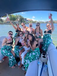 Eight women in matching green leaf sarongs and black swim tops sipping drinks and posing on a sunny party boat in a palm‑lined waterfront canal with mansions in the background.