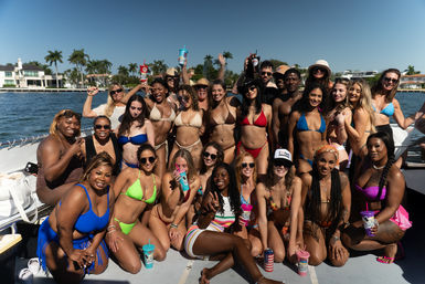 Large group of people in colorful bikinis and swimwear smiling and posing on a sunny boat party, holding drinks with palm trees and waterfront homes in the background