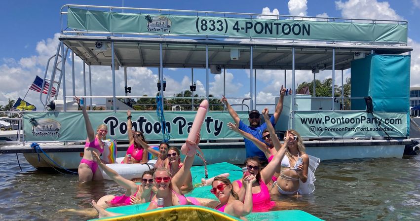 Group of friends in bright pink swimsuits cheering on floating mats in shallow South Florida water in front of a double-deck pontoon party boat under a sunny blue sky
