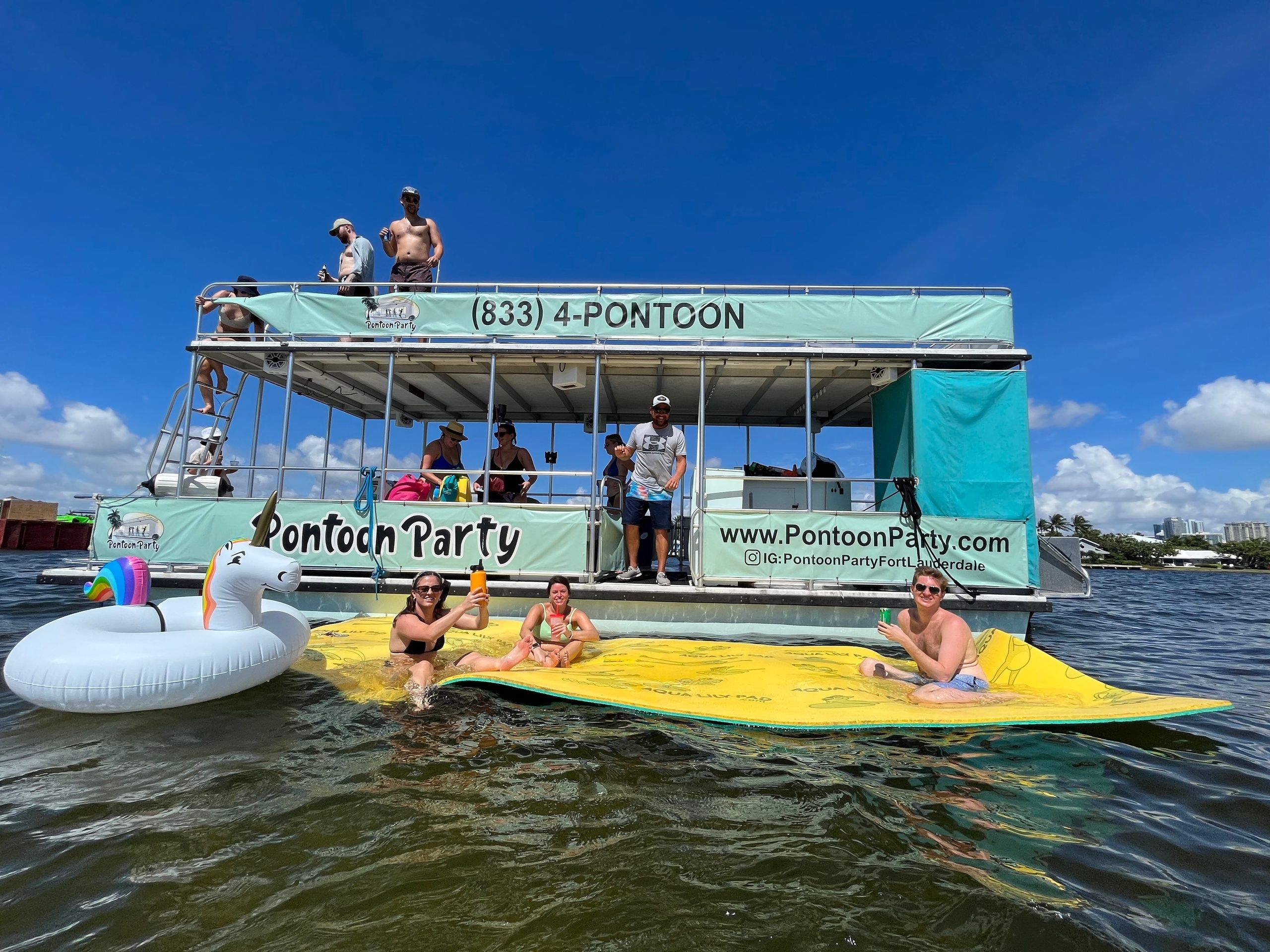 Group enjoying a sunny pontoon boat party in Fort Lauderdale, Florida — lounging on a large yellow floating mat and unicorn inflatable on calm coastal water under a clear blue sky.