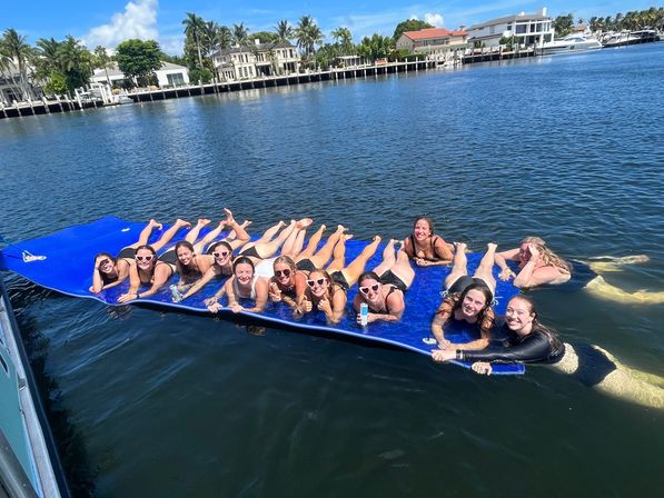 Group of friends lounging on a large blue floating mat in a sunny waterfront canal, smiling and holding drinks with docks, boats, palm trees and waterfront homes along the shoreline