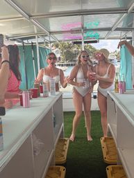 Three women in white swimsuits laughing and holding drinks on a pontoon boat party, neon “Pontoon Party” sign overhead and palm-tree lined waterfront with boats in the background.