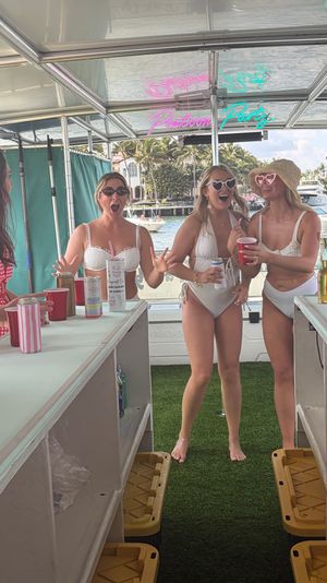 Three women in white swimsuits laughing and holding drinks on a pontoon boat party, neon “Pontoon Party” sign overhead and palm-tree lined waterfront with boats in the background.