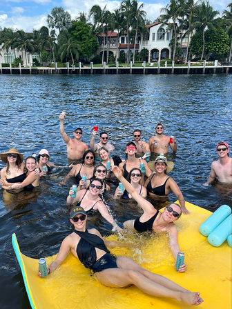 Smiling group of people in swimsuits partying on a large yellow float in a sunny palm-lined waterfront canal, holding drinks with luxury waterfront homes in the background.