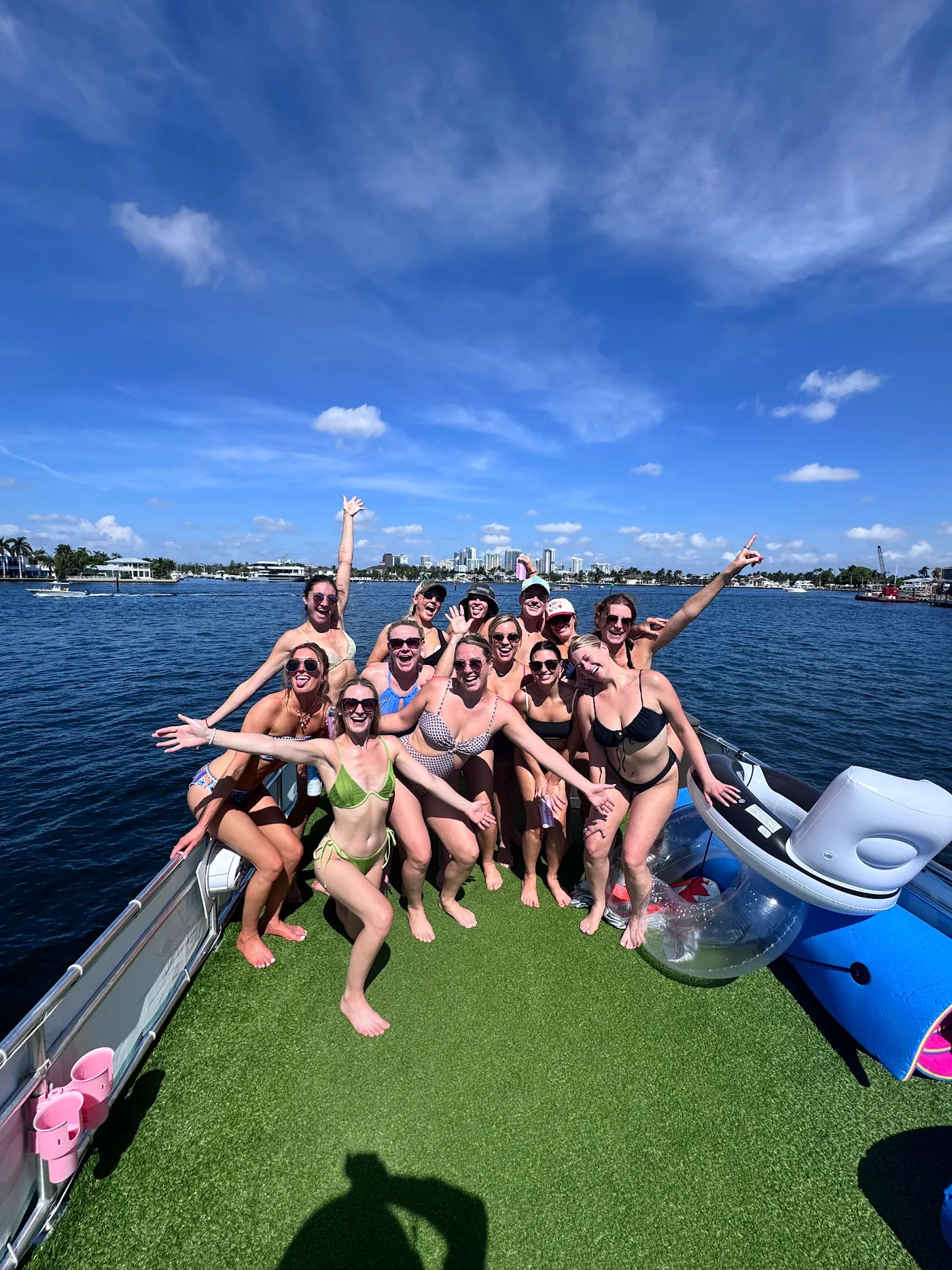 Boat party with a cheerful group of women in bikinis posing on a pontoon near a waterfront city skyline under a bright blue sky