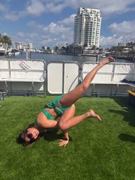 Person in green swimsuit and sunglasses holding a one-arm yoga/acrobatic balance on artificial turf on a boat deck, with yachts, marina and high-rise waterfront condos under a sunny blue sky.