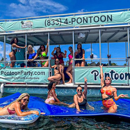 Group of women in colorful swimsuits partying on a pontoon boat and floating mat, cheering with drinks under a bright blue sky on calm coastal waters.