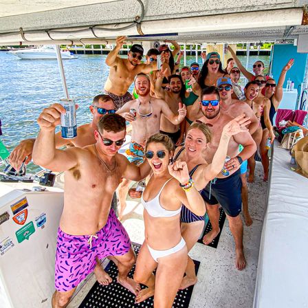 Group of young adults in swimsuits partying on a covered boat near a marina, cheering with drinks and smiling on a sunny summer day
