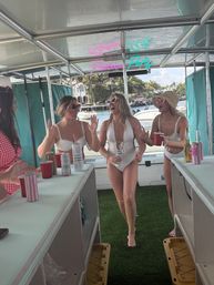 Four women laughing on a pontoon boat at a sunny waterfront, wearing swimsuits and sunglasses, holding drinks under a neon “Pontoon Party” sign with palm trees and yachts in the background.