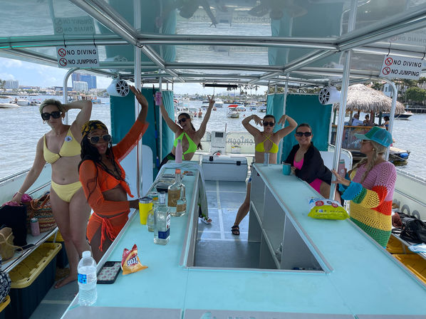 Sunny waterfront party on a covered pontoon boat — group of women in colorful swimsuits and cover-ups posing around a central bar with drinks and snacks, anchored boats and a marina in the background.
