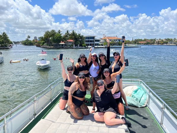 Boat party: group of women in black swimsuits and caps cheering with drinks on a sunny day along a palm‑lined waterfront canal with luxury homes.
