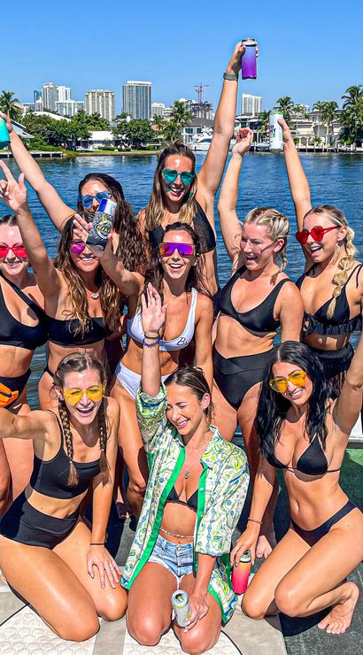 Boat party with a group of friends in bikinis raising colorful drinks and wearing sunglasses on a sunny waterfront with palm trees and a city skyline.