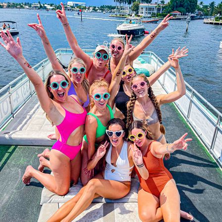 Group of friends on a sunny pontoon boat party wearing colorful swimsuits and playful flower-shaped sunglasses, cheering near a palm-lined marina with yachts in the background.