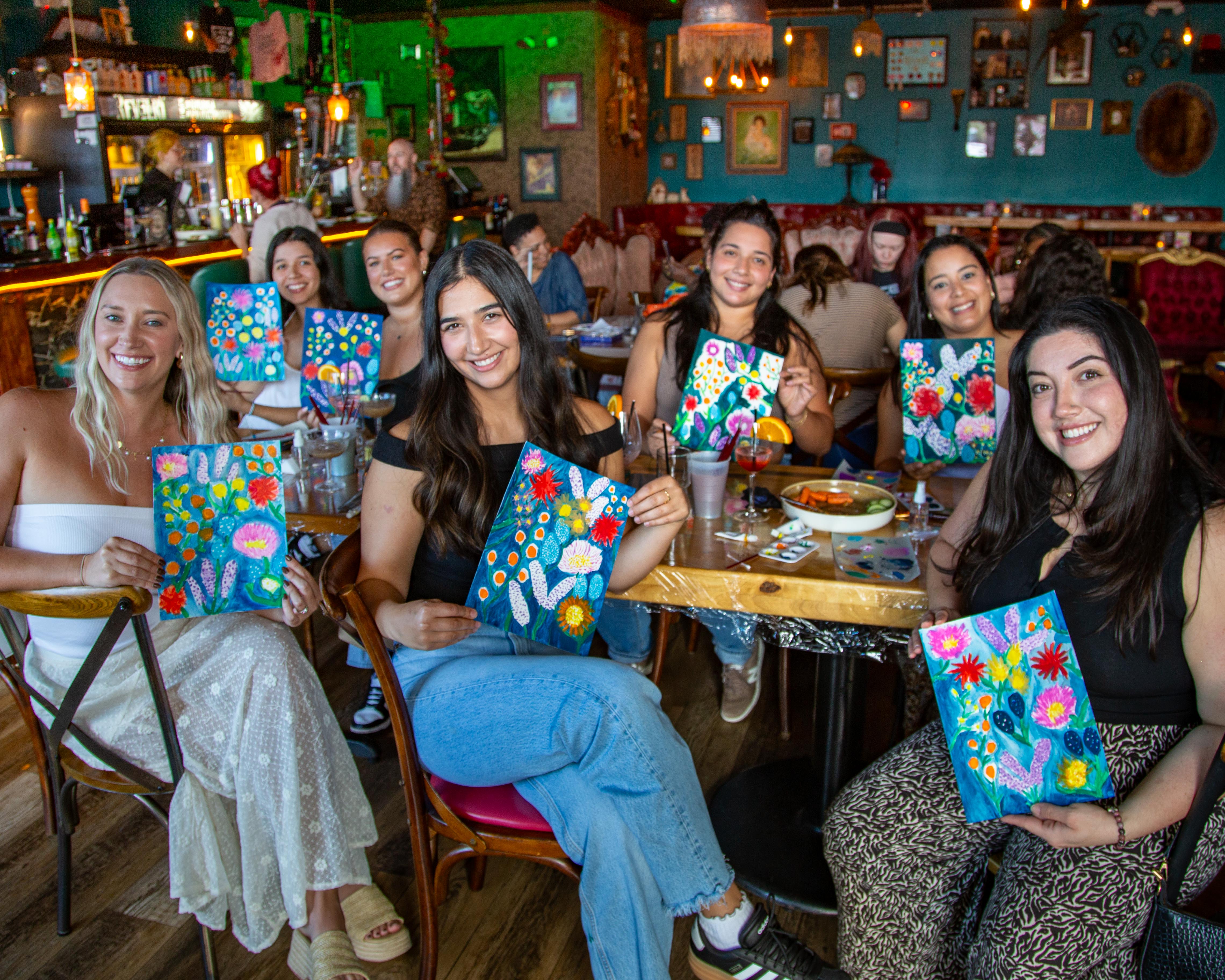Group of friends at a lively paint-and-sip in a cozy cafe, smiling and holding colorful floral canvases around a wooden table with drinks and art supplies