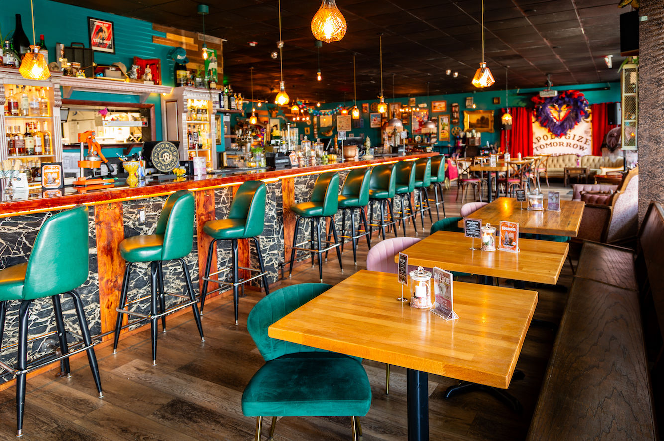 Vibrant retro cocktail bar interior with emerald green barstools along a long wooden counter, warm pendant lights, and cozy dining tables.