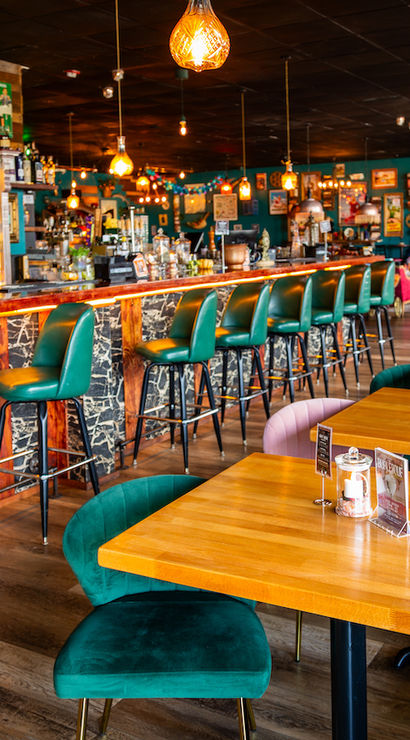 Vibrant retro cocktail bar interior with emerald green barstools along a long wooden counter, warm pendant lights, and cozy dining tables.