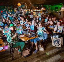 Lively paint-and-sip party at a cozy restaurant bar with a large group seated at long tables proudly holding colorful floral canvases during a social group painting workshop.