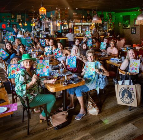 Lively paint-and-sip party at a cozy restaurant bar with a large group seated at long tables proudly holding colorful floral canvases during a social group painting workshop.