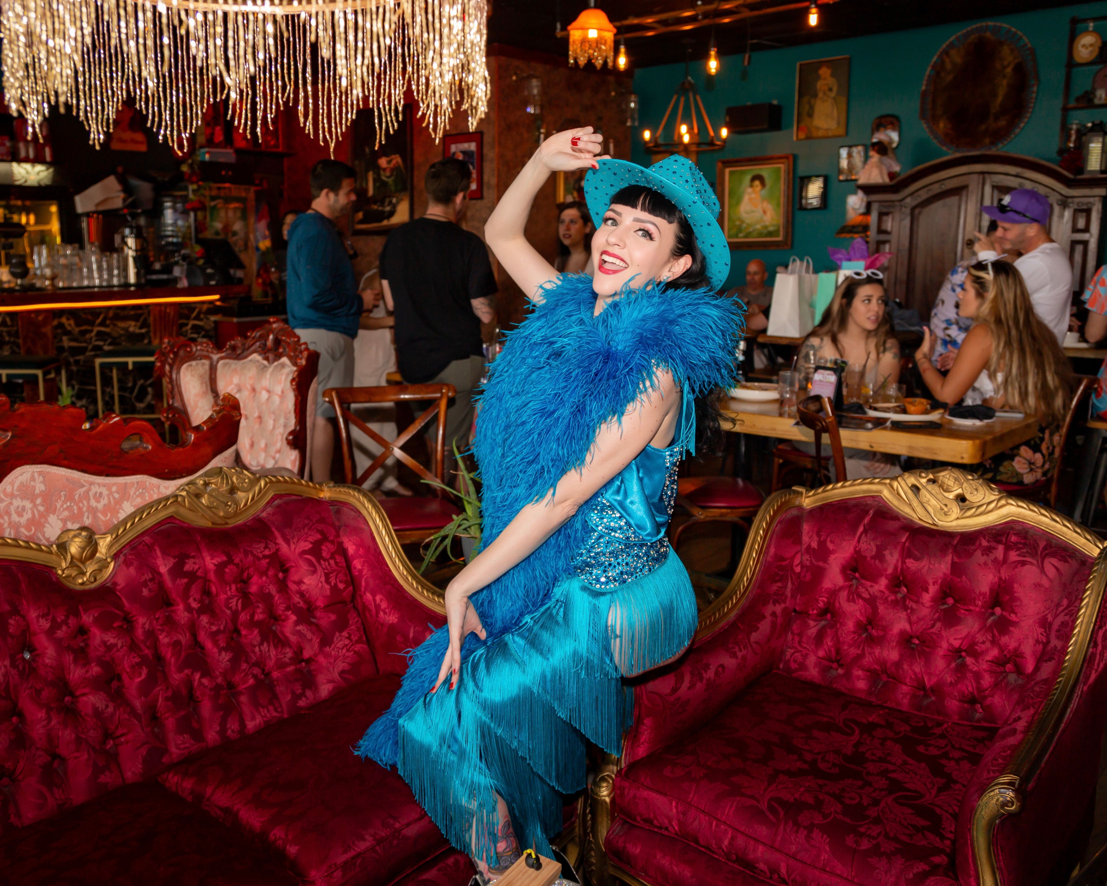 Flamboyant performer in a turquoise fringe dress, feather boa and hat striking a pose on a red velvet antique sofa inside a colorful cabaret-style lounge with chandeliers and diners.