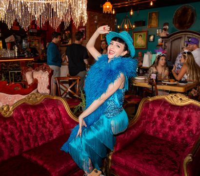 Flamboyant performer in a turquoise fringe dress, feather boa and hat striking a pose on a red velvet antique sofa inside a colorful cabaret-style lounge with chandeliers and diners.