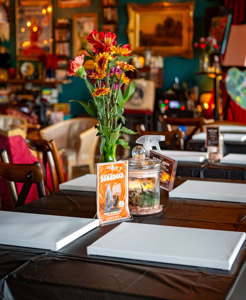 Cozy indoor table setup with blank canvases, a vase of vibrant red and yellow flowers, glass terrarium and event flyer ready for a paint-and-sip or community art workshop.