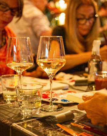 Two wine glasses and a water glass on a table at a lively paint-and-sip night, paint palette, brushes and blurred people in the background.
