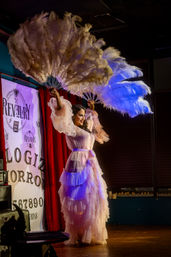 Cabaret performer striking a dramatic pose on an indoor stage in a layered pink ruffled gown, holding oversized white feather fans aloft under colorful theatrical lighting.