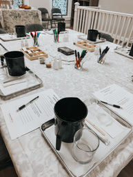 Candle-making workshop table set up with metal trays, black pouring pitchers, glass jars, stirring tools, small bottles, colored pens and printed worksheets in a bright craft studio