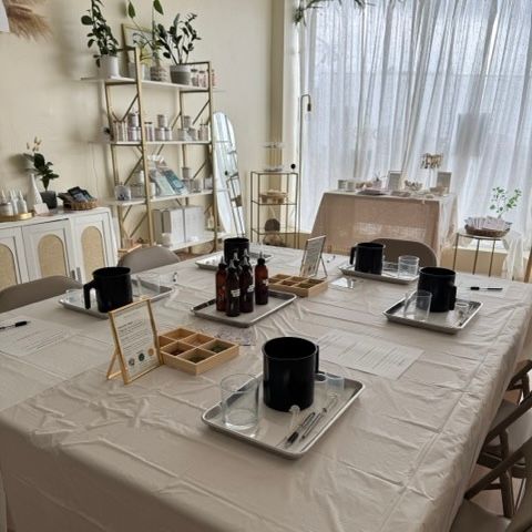 Sunlit DIY candle-making workshop setup on a large table with metal trays, black pouring pots, glass beakers, amber bottles, wooden sample box and instruction sheets in a cozy bright studio with shelves, plants and sheer curtains.
