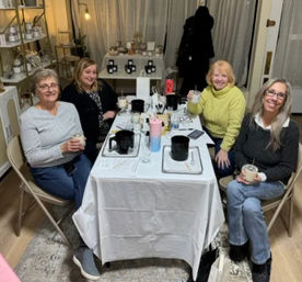 Four women smiling around a table at a cozy craft studio workshop, holding drinks and working with candle-making pots, jars, tools and instruction sheets, with shelves and warm lighting in the background.