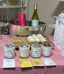 Indoor celebration table with chilled wine bottles in a metal ice bucket, a tray of sprinkled mini cupcakes, four labeled scented candles topped with dried botanicals, inspirational cards and white gift bags on a pink tablecloth.