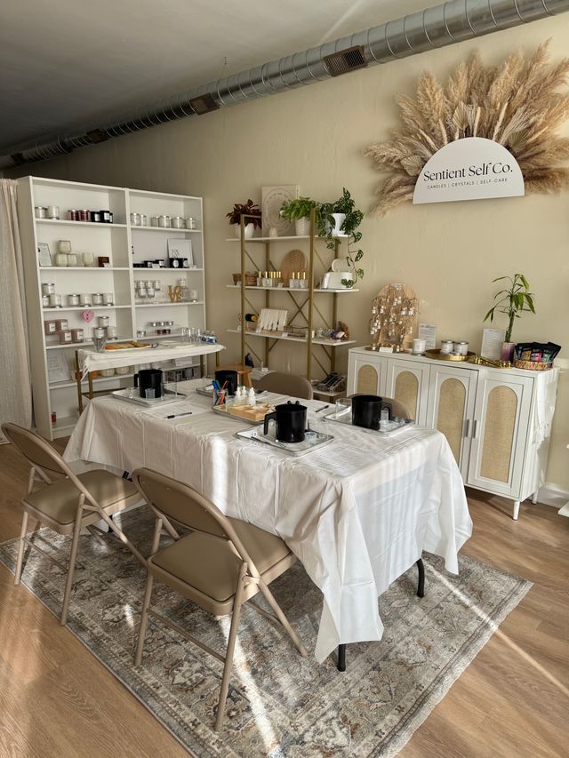 Sunlit, neutral-toned studio set for a candle-making workshop — folding chairs around tables with white linens and black pouring pots, shelves of jars and candles, pampas grass wall decor and potted plants.
