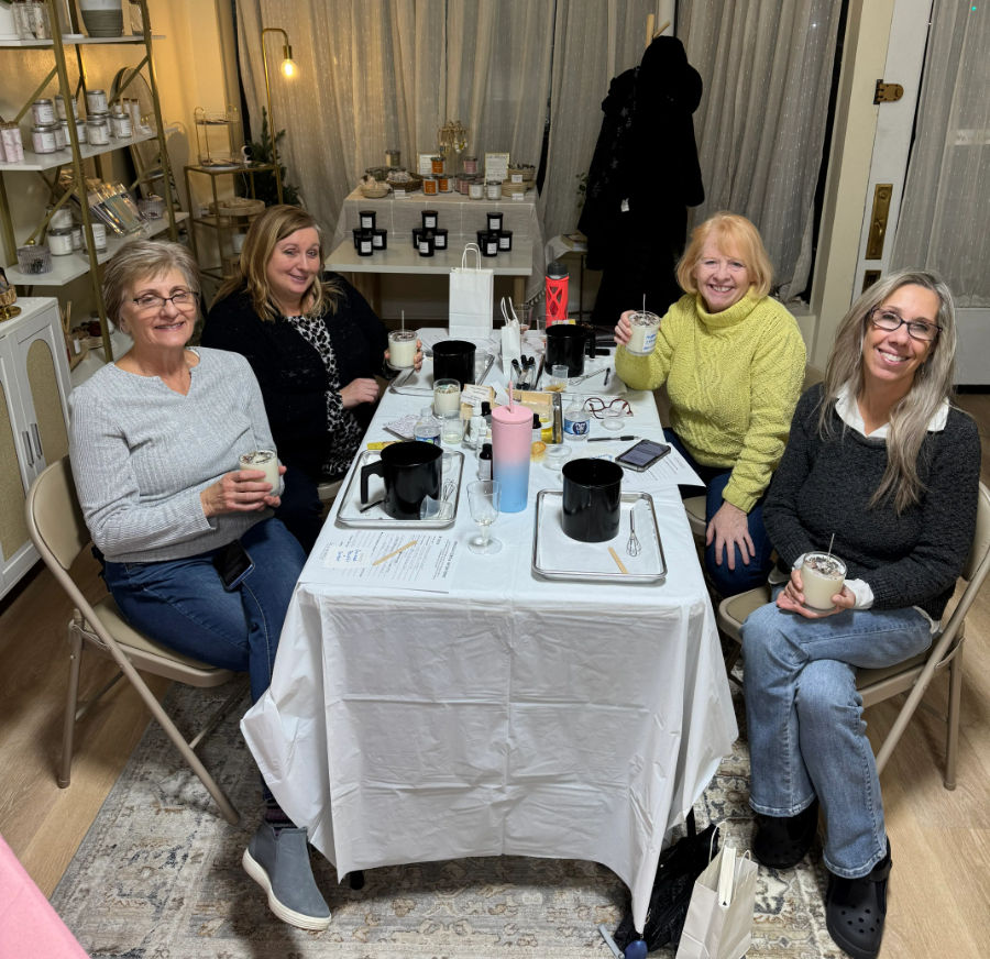 Four smiling women seated around a cozy indoor table at a candle-making workshop, holding jar candles amid pouring pots, supplies and tasting glasses.