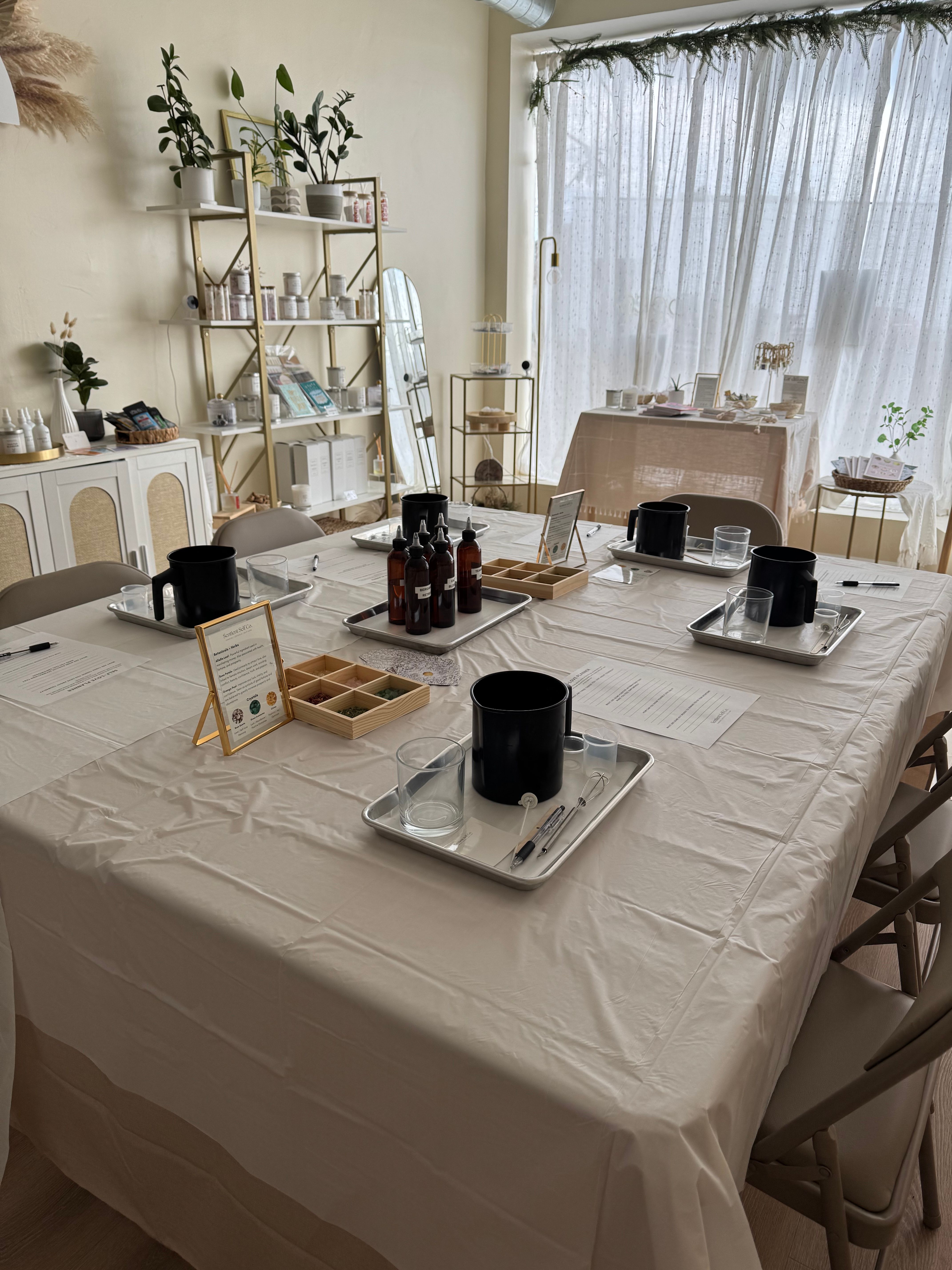 Sunlit craft studio set for a DIY candle-making workshop with a large table, trays holding black pouring pots, glass beakers, amber fragrance bottles, wooden scent sample box, and plant-filled shelving against sheer curtains.