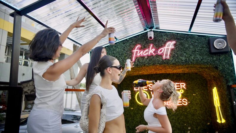 Four women in white outfits partying on an urban rooftop patio, raising cans and pouring champagne under a clear canopy in front of a green wall with a pink neon 'fuck off' sign