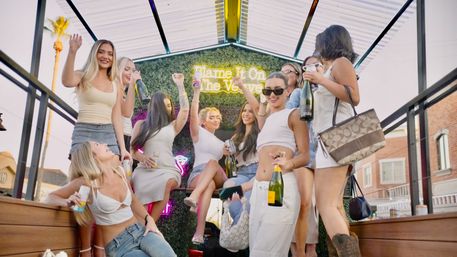 Group of young women celebrating on a sunny urban rooftop patio, wearing summer outfits and sunglasses, holding champagne bottles and canned drinks, smiling and cheering in front of a neon-lit green wall with palm tree and brick buildings visible.